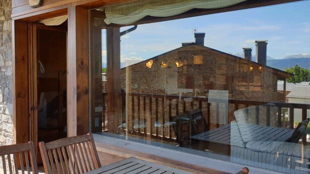 A wooden balcony with a stone wall, a glass door, and a view of a stone building with chimneys.