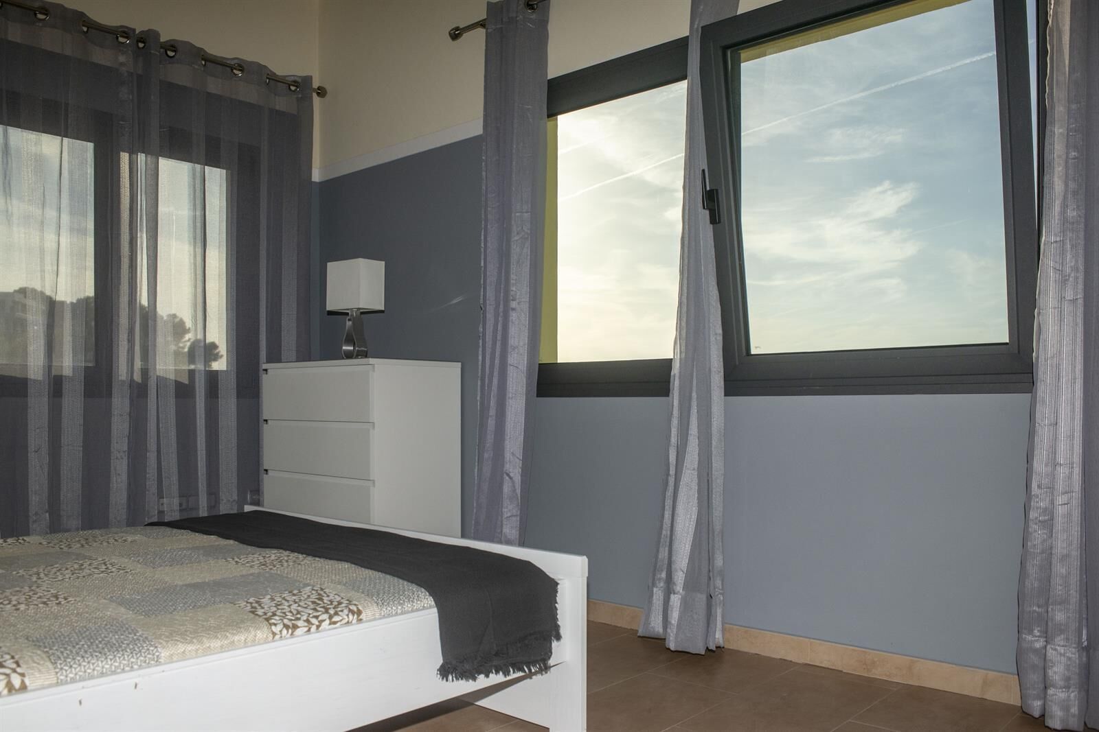 Une chambre à coucher avec un lit, une commode, une fenêtre avec des rideaux et une vue sur le ciel.