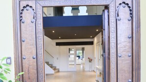 Une porte double en bois, ornée de sculptures complexes, donne sur un couloir où se trouve une moquette bleue et un escalier.