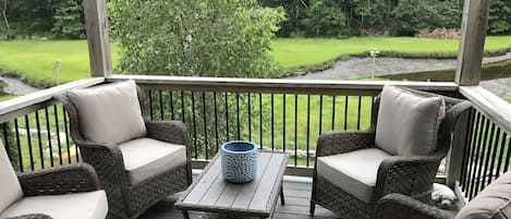 A covered outdoor seating area with wicker furniture and a table, overlooking a grassy landscape and trees.