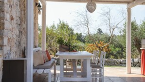 A covered outdoor seating area with a table and chairs, a stone wall, and a view of trees and sky. - TD Trulli della Lama Private Trulli with Pool (Castellana Grotte)