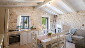 A rustic kitchen with stone walls, wooden beams, a dining table with chairs, a stove, and a white cabinet. - TD Trulli della Lama Private Trulli with Pool (Castellana Grotte)