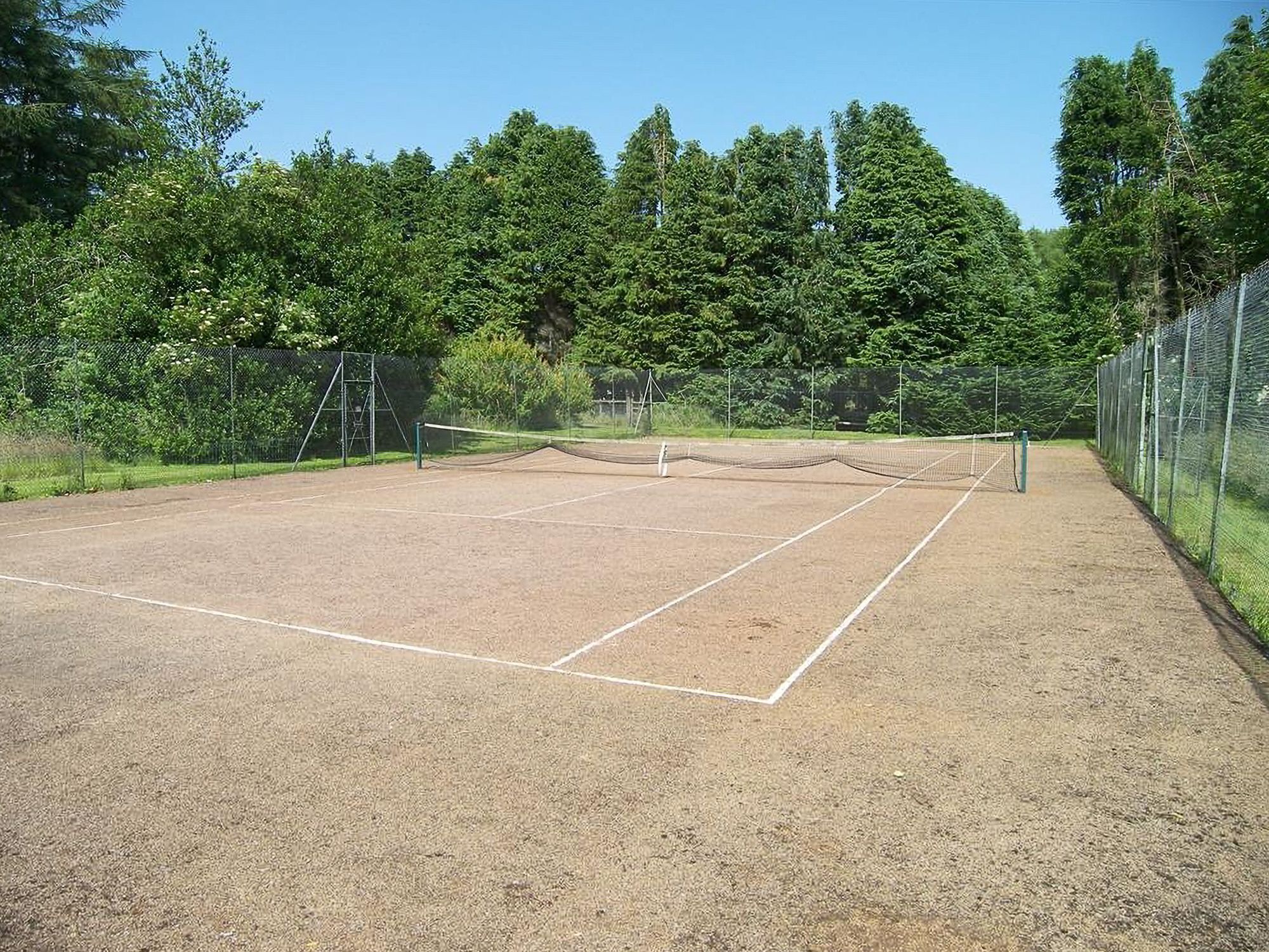 Un court de tennis extérieur, doté d’un filet, entouré d’une clôture et d’arbres.