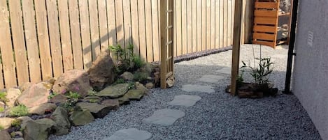 A wooden gate leading to a gravel path with stepping stones, surrounded by a wooden fence and a small garden area with rocks and plants.