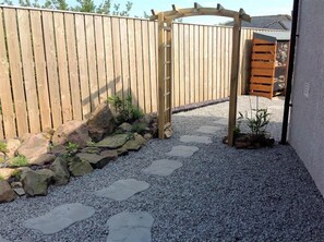 A wooden gate leading to a gravel path with stepping stones, surrounded by a wooden fence and a small garden area with rocks and plants.