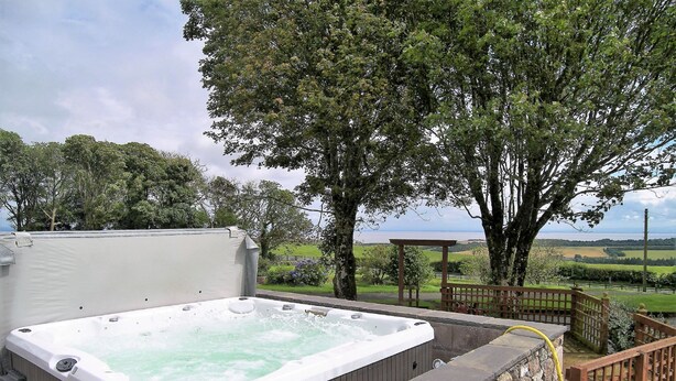 Un bain à remous doté d’une terrasse en bois et d’un mur en pierre, entouré d’arbres et offrant une vue sur la campagne.