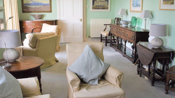 Een woonkamer met een beige bank, een witte fauteuil en een witte salontafel. Er staan twee lampen op bijzettafeltjes, een houten kast en een deur op de achtergrond.