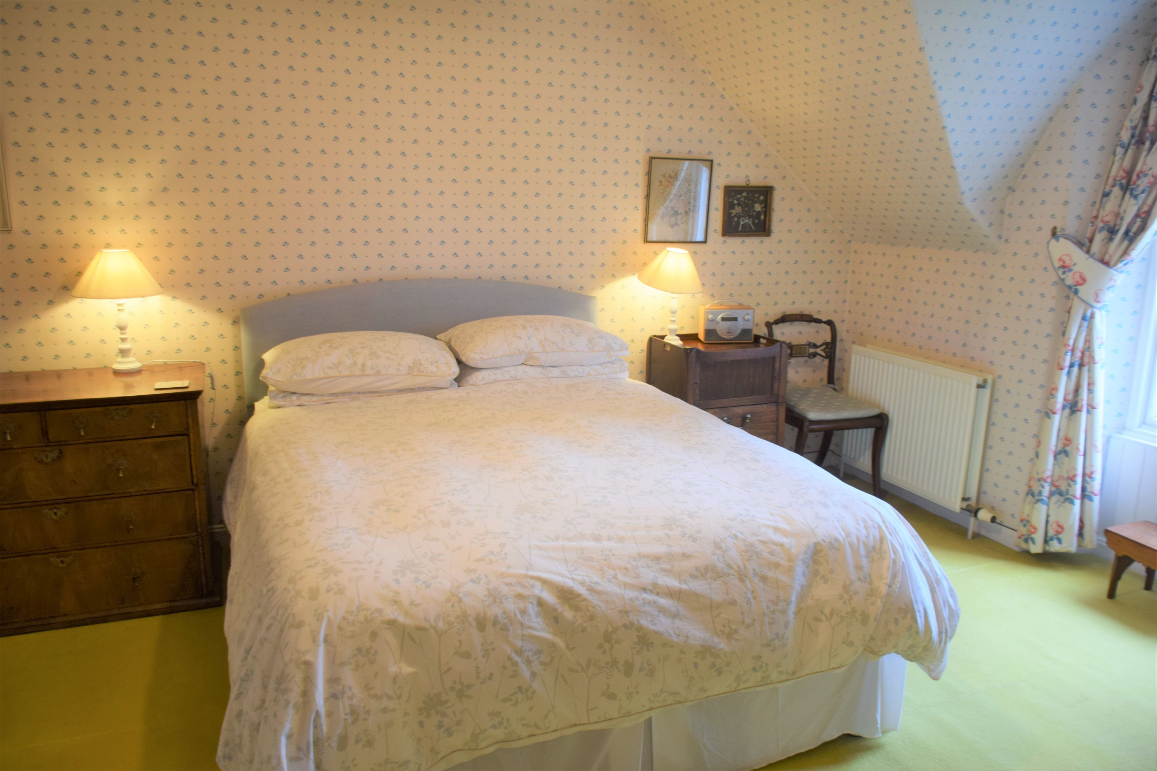 A bedroom with a bed, two bedside lamps, a dresser, a chair, and a radiator.