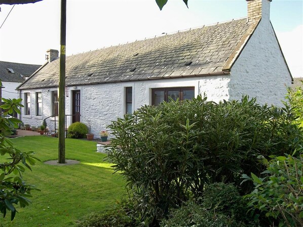 A white stone house with a sloped roof, a front door, and a small front yard with a bush.