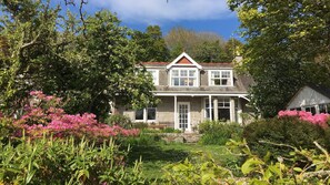 Une maison à deux étages avec un porche à l’avant, entourée d’une végétation luxuriante et d’arbustes en fleurs.