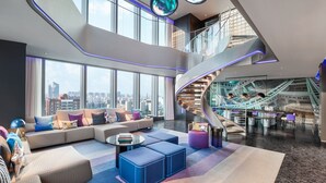 A spacious lobby with a curved staircase, large windows offering city views, and a seating area with sofas and ottomans.