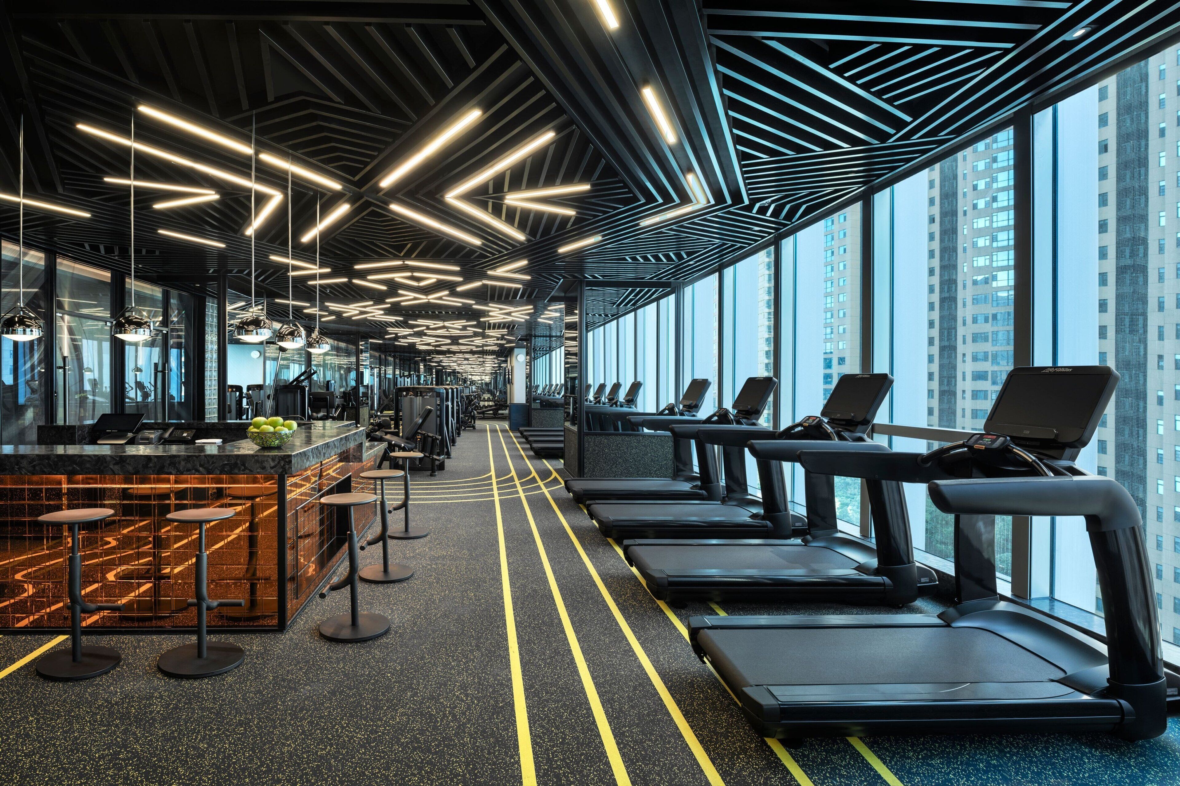 A modern gym with treadmills, a bar area, and large windows offering city views.
