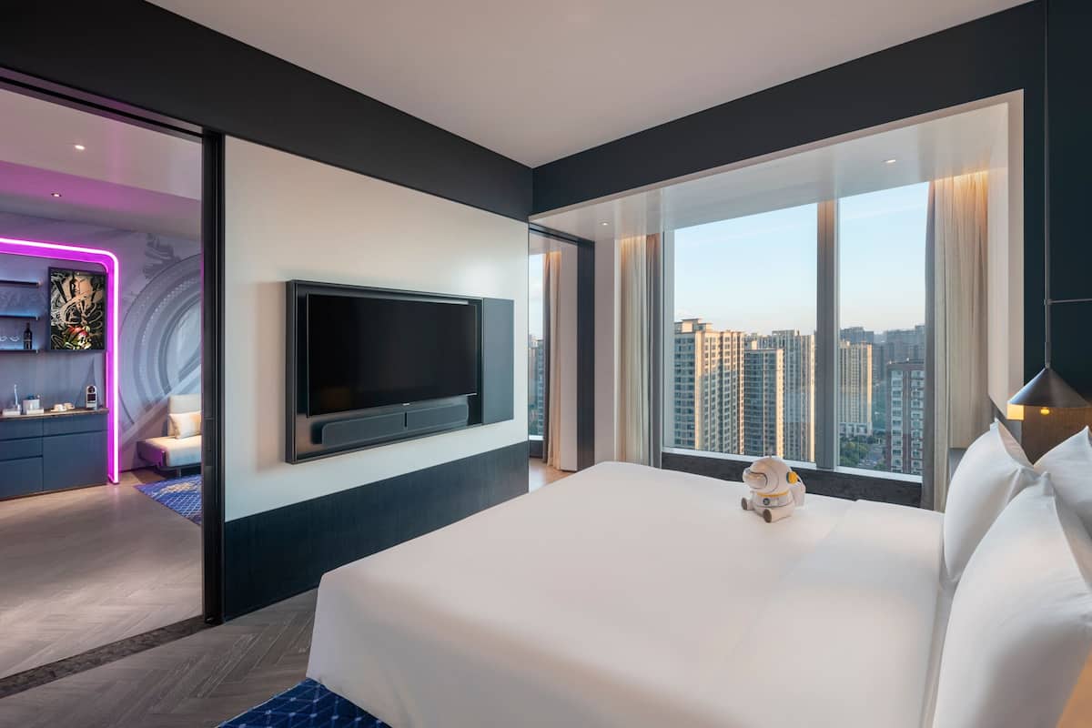 Ein modernes Hotelzimmer mit einem großen Bett, einem flachbildfernseher an der Wand und Blick auf die Stadt durch große Fenster.