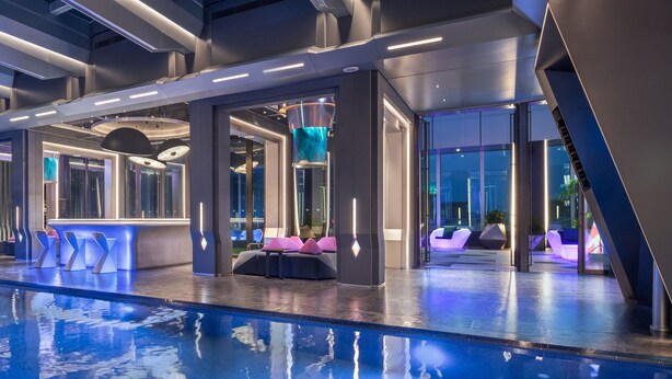 A modern indoor pool area with a clear pool, seating areas with cushions, and a bar.