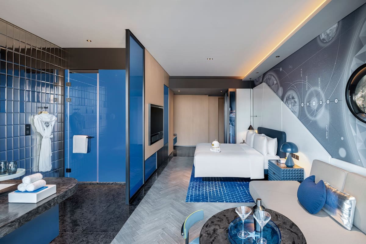 Ein modernes Hotelzimmer mit einem großen Bett, einer Sitzecke mit Tisch, einem Fernseher und einem Badezimmer mit Spiegel und Handtuchhalter.