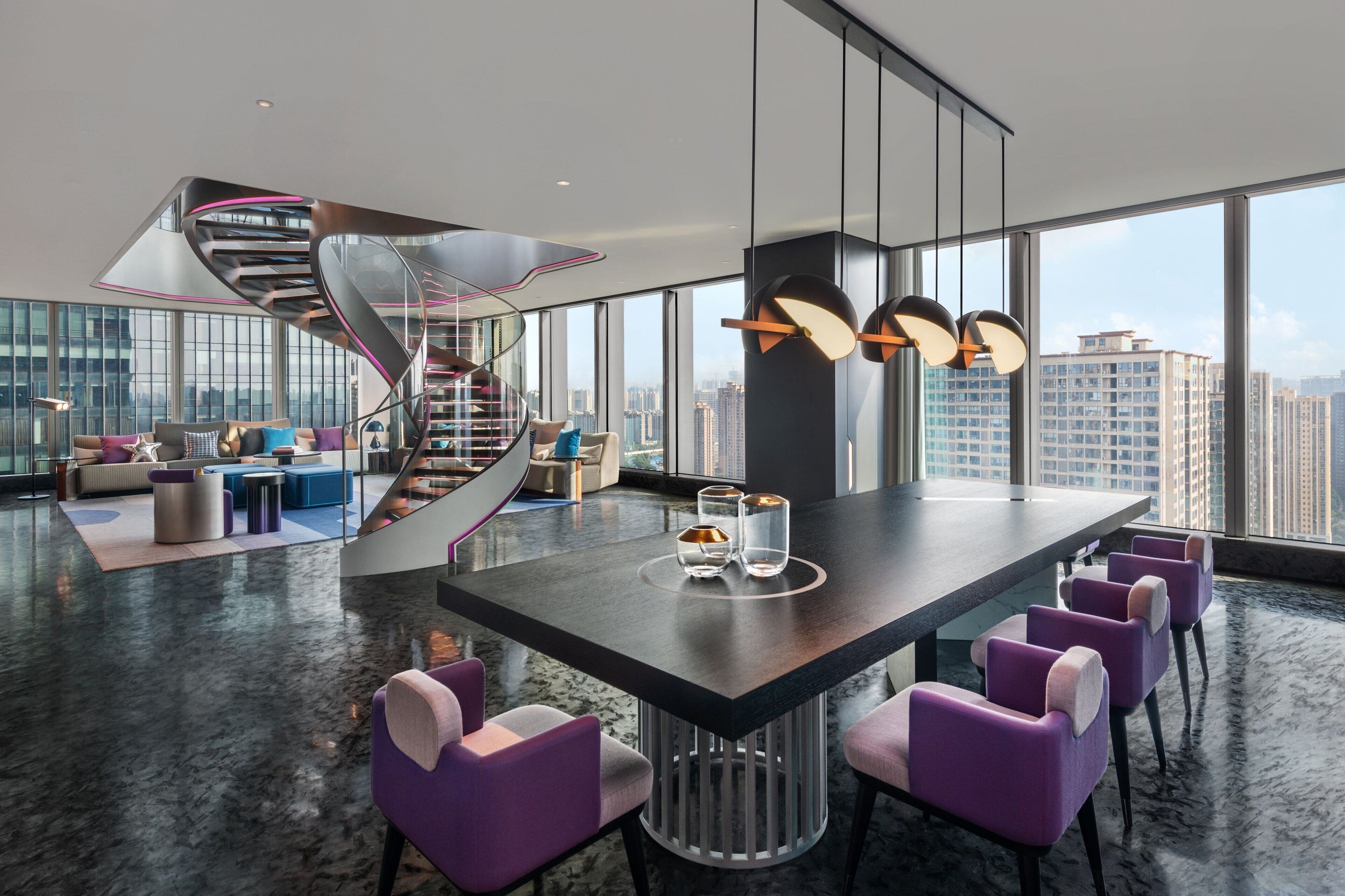 A modern interior with a central dining table, purple chairs, a spiral staircase, and large windows offering city views.