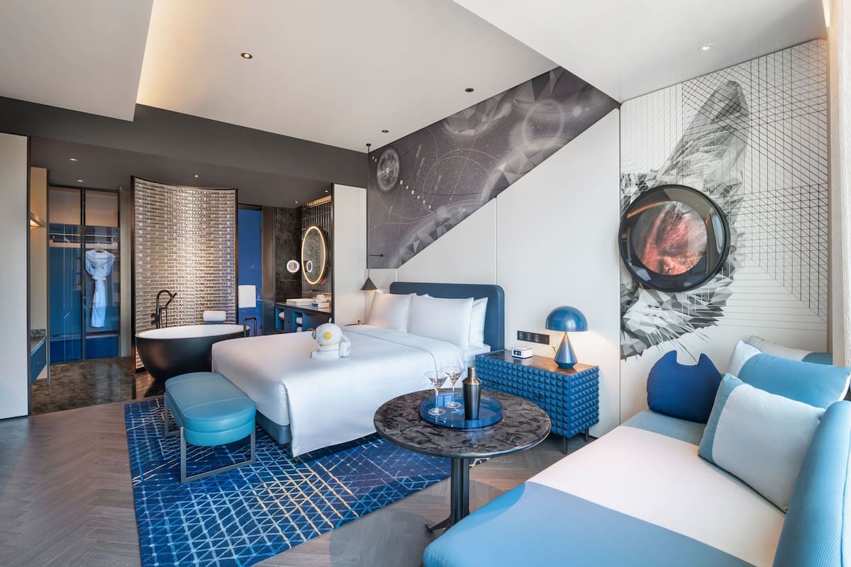 Ein modernes Hotelzimmer mit einem großen Bett, einer Sitzecke, einer Badewanne und einer Wand mit einem Wandgemälde zum Thema Weltall.