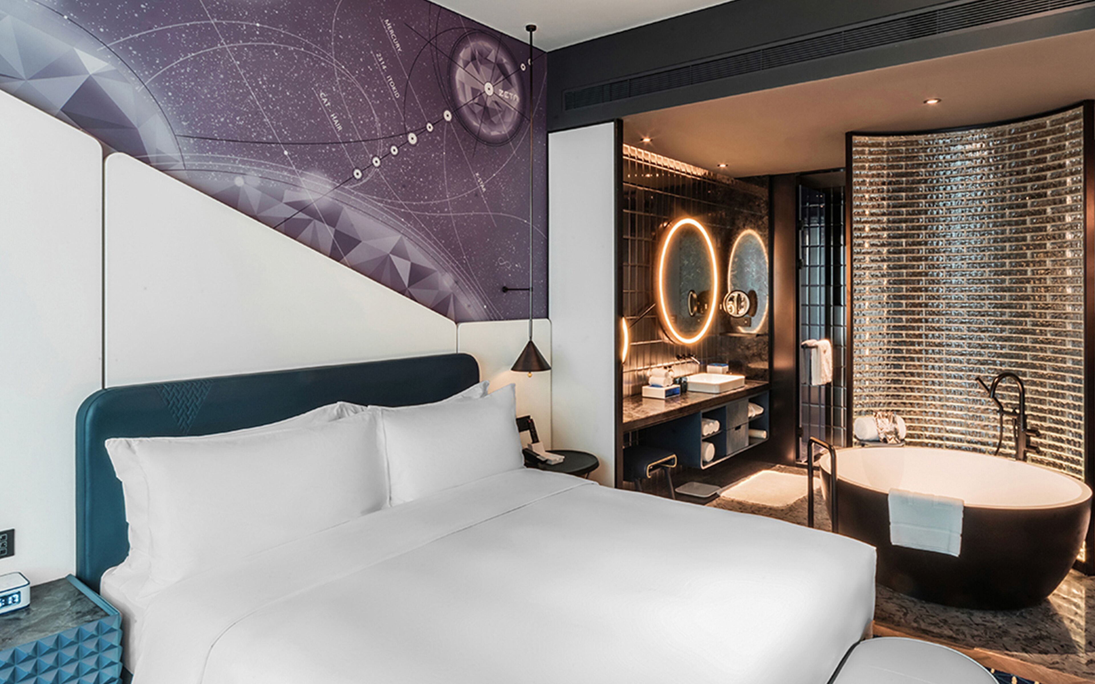 A modern hotel room with a large bed, a vanity with a mirror, and a bathtub.