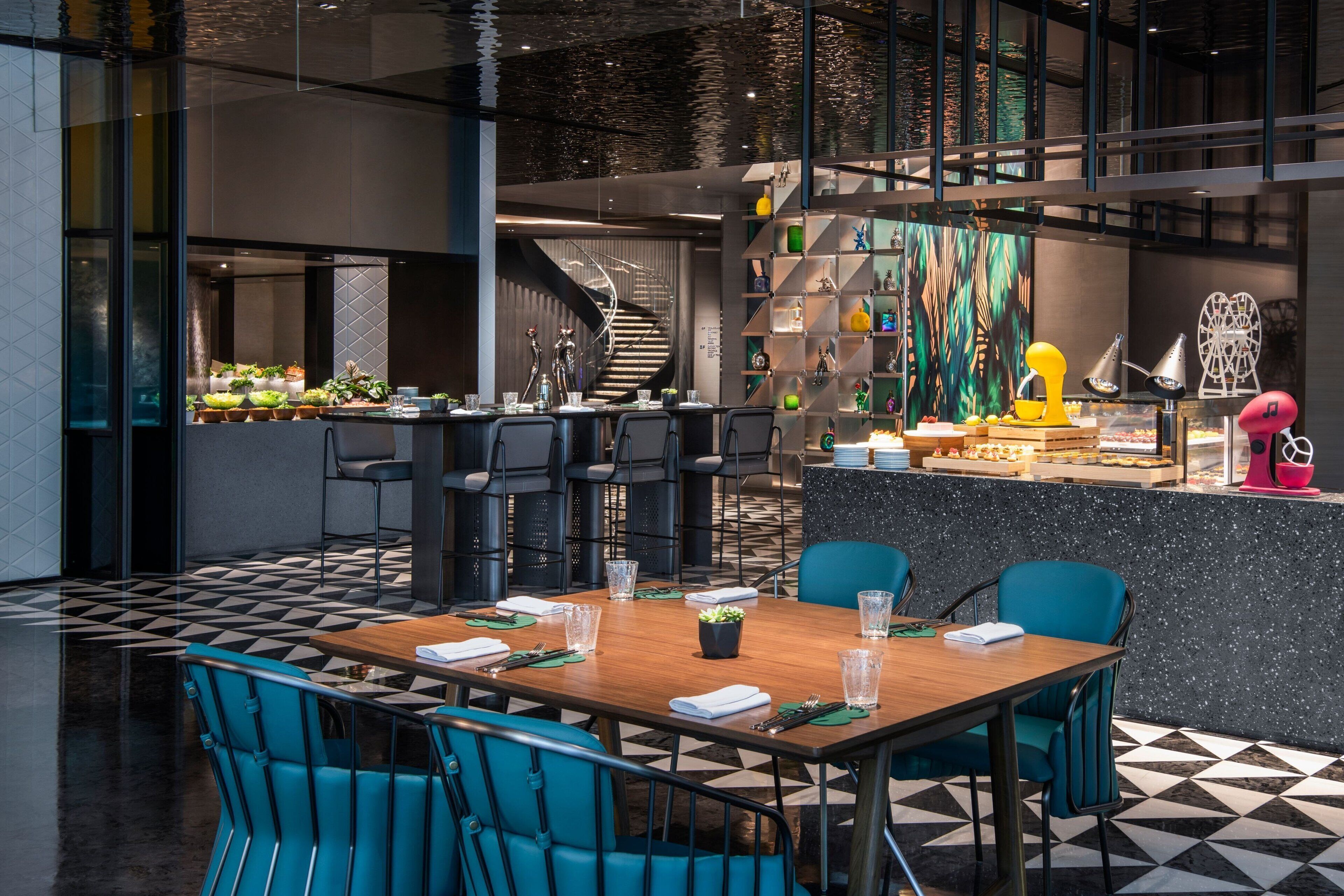 A modern restaurant with a central wooden table, blue chairs, and a black and white checkered floor.