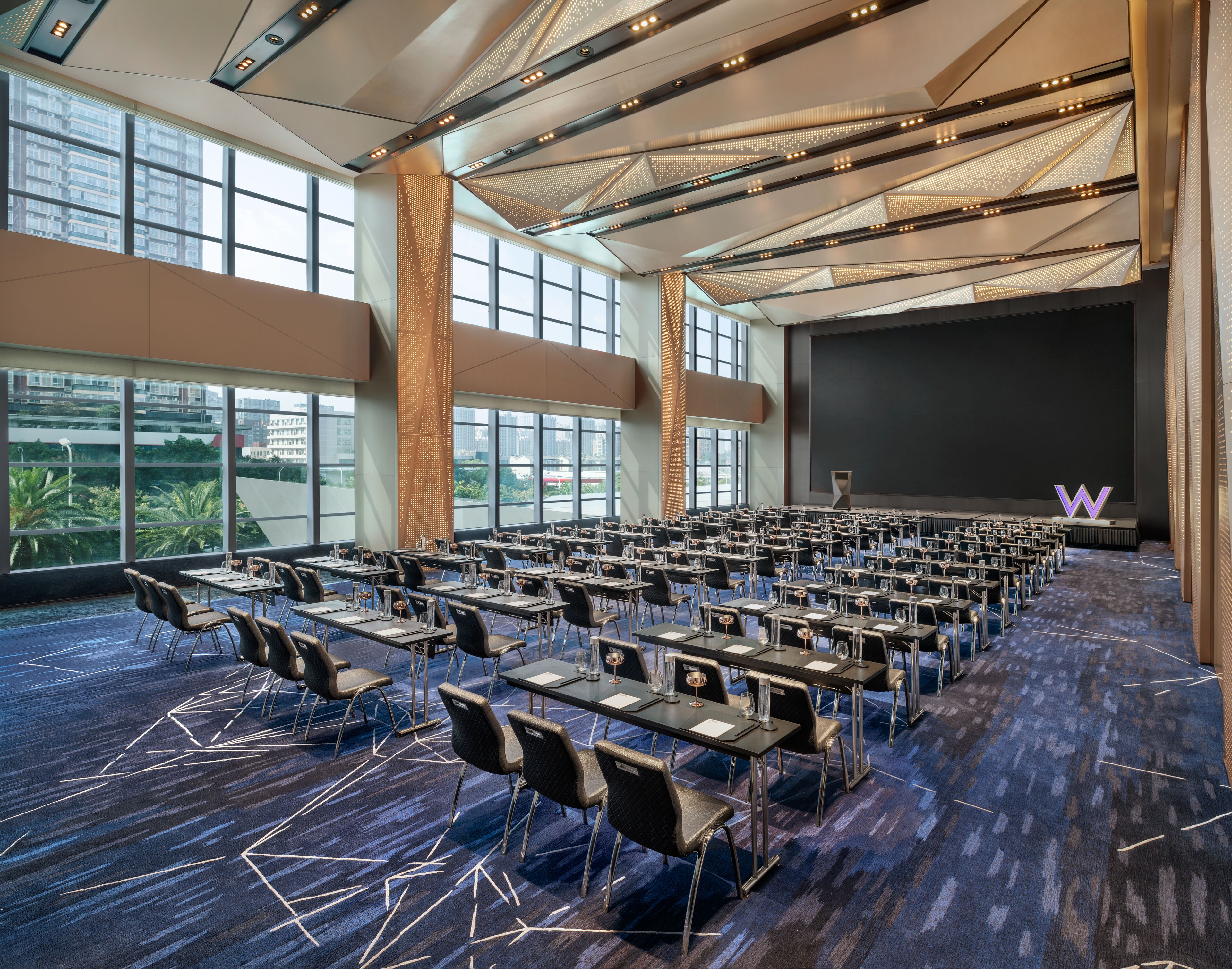 A conference room with rows of tables and chairs, large windows, and a stage with a screen.