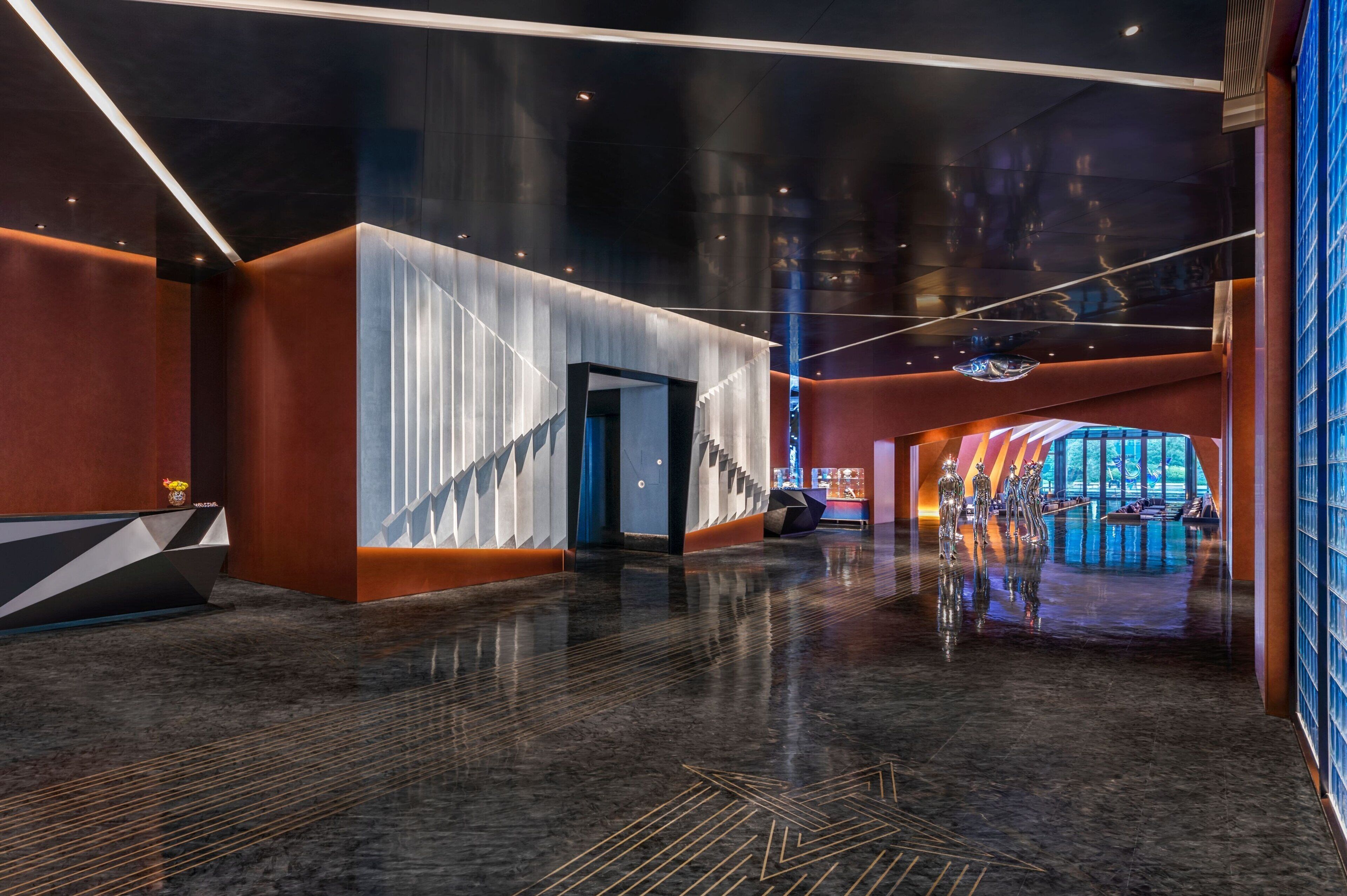 A spacious lobby with a modern design, featuring a central staircase, a reception area, and a large window offering a view of the outdoors.