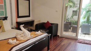 A hotel room with a bed, a desk, a chair, a sofa, and a sliding glass door leading to an outdoor area with plants. - Citrus Hotels Sriperumbudur (Sriperumbudur)