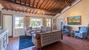 A spacious living room with a high wooden ceiling, large windows, a sofa, armchairs, and a painting on the wall.
