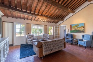 Uma sala de estar ampla com teto de madeira alto, janelas grandes, um sofá, poltronas e um quadro na parede.