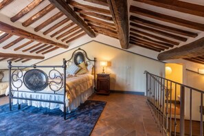 Quarto com teto de madeira, cama com estrutura de metal, mesa de cabeceira e escada com corrimão.