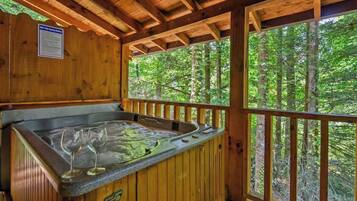 A wooden cabin with a hot tub, wooden deck, and a view of a forest.