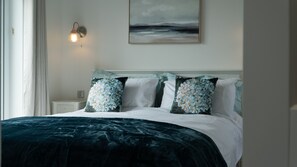 A neatly made bed with floral pillows, a wall-mounted light, and a painting above it. - Beach House Paradise Found WF-Fi, Netflix, Pool,Gym, Pet Friendly close to beach (Filey)