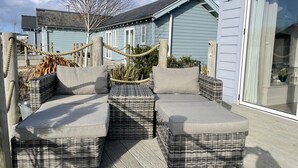 A wicker sofa set with cushions on a wooden deck, overlooking a house with a blue exterior.
