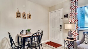 A dining area with a table and chairs, a small side table, a red doormat, and a wall-mounted sign that reads "EAT".