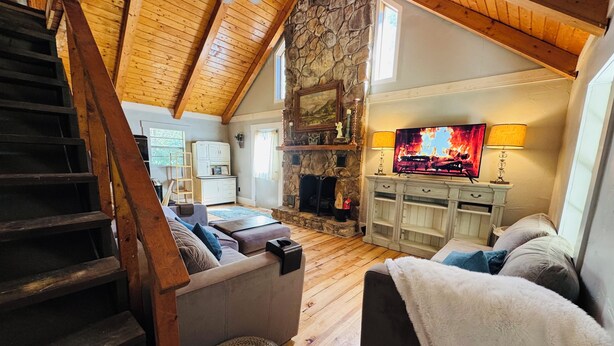 A cozy living room with a stone fireplace, wooden beams, a TV, and a staircase. - Waterfront Cabin on trophy trout stream, fire pit, smart tv, fireplace (Ellijay)