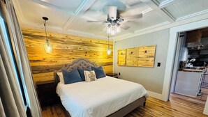 A bedroom with a wooden headboard, a bed with white bedding, a ceiling fan, and a small kitchen area visible through an open door. - Waterfront Cabin on trophy trout stream, fire pit, smart tv, fireplace (Ellijay)