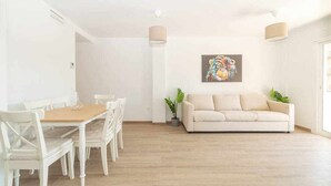 Un salon lumineux avec une table à manger en bois, des chaises blanches, un canapé beige et un tableau coloré accroché au mur.