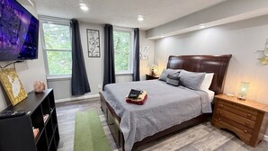 Ein Schlafzimmer mit Bett, Nachttischen, Fernseher, Bücherregal und einem Fenster mit Vorhängen.