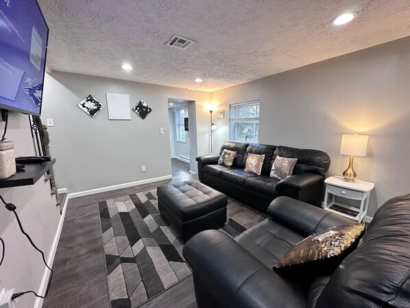 A living room with a black leather sofa, a matching ottoman, a small white side table, and a TV on a stand. - 100% renovated House 8 minutes to downtown (Pittsburgh)