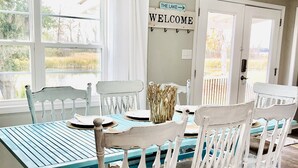 Un coin repas avec une table de couleur turquoise et des chaises blanches, un panneau indiquant « BIENVENUE », et une vue sur l’extérieur grâce à de grandes fenêtres.