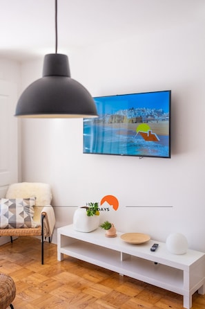 En moderne stue med en hvid sofa, et hvidt konsolbord, en sort hængelampe og et fladskærms-tv på væggen, der viser en feriereklame.