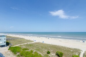 A beach with a building in the foreground, sandy beach, and clear blue sky. - Premier Oceanfront Condo w Rooftop Deck (Carolina Beach)
