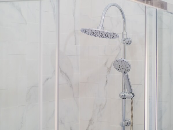 Ducha con cabezal de ducha manual y cabezal de ducha de lluvia fijos, ambos con acabados cromados, montados en una pared con azulejos de mármol.