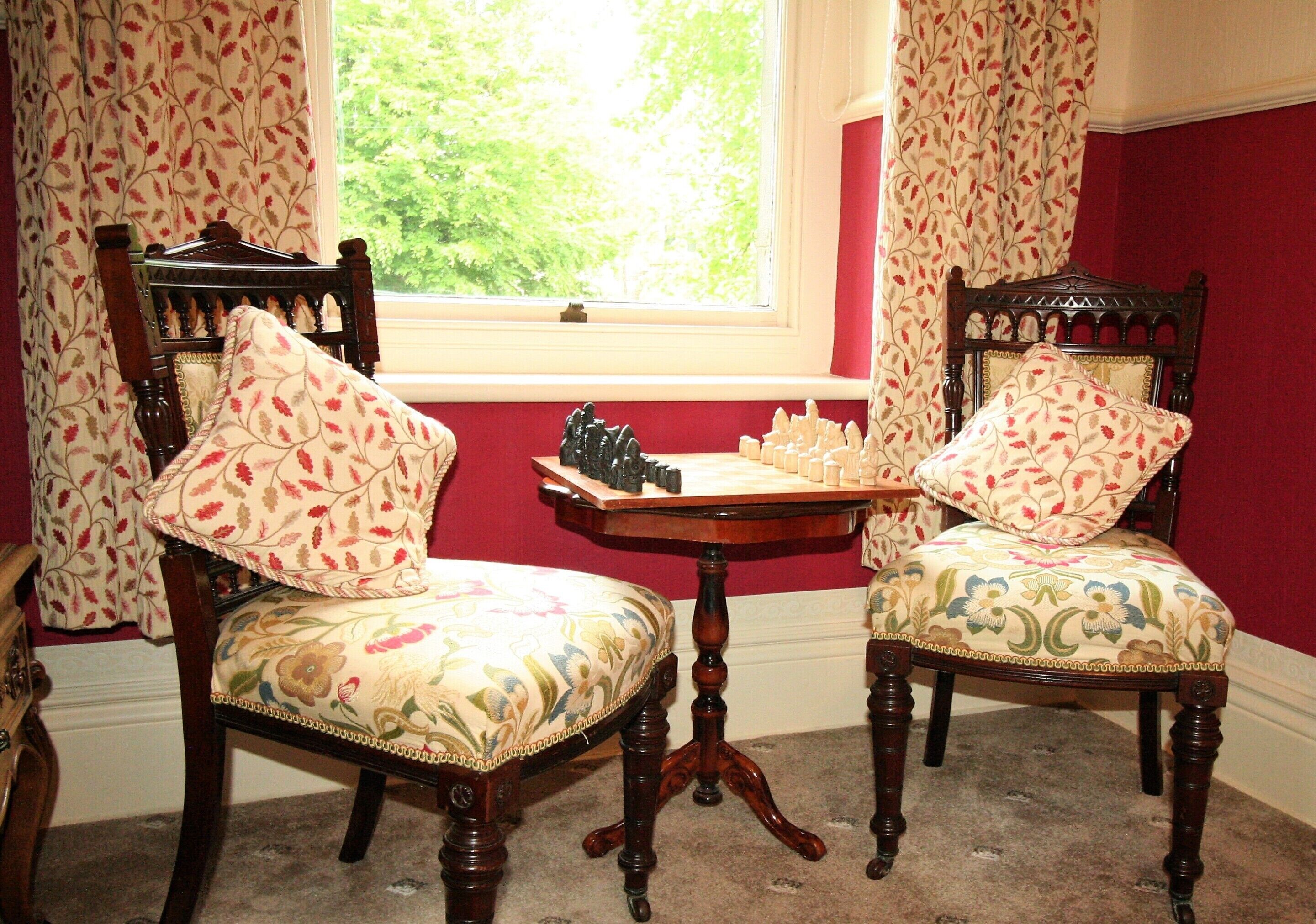 Deux fauteuils en bois richement décorés, recouverts d’un tissu à motifs floraux, et un jeu d’échecs posé sur une petite table, le tout placé près d’une fenêtre donnant sur un paysage verdoyant.