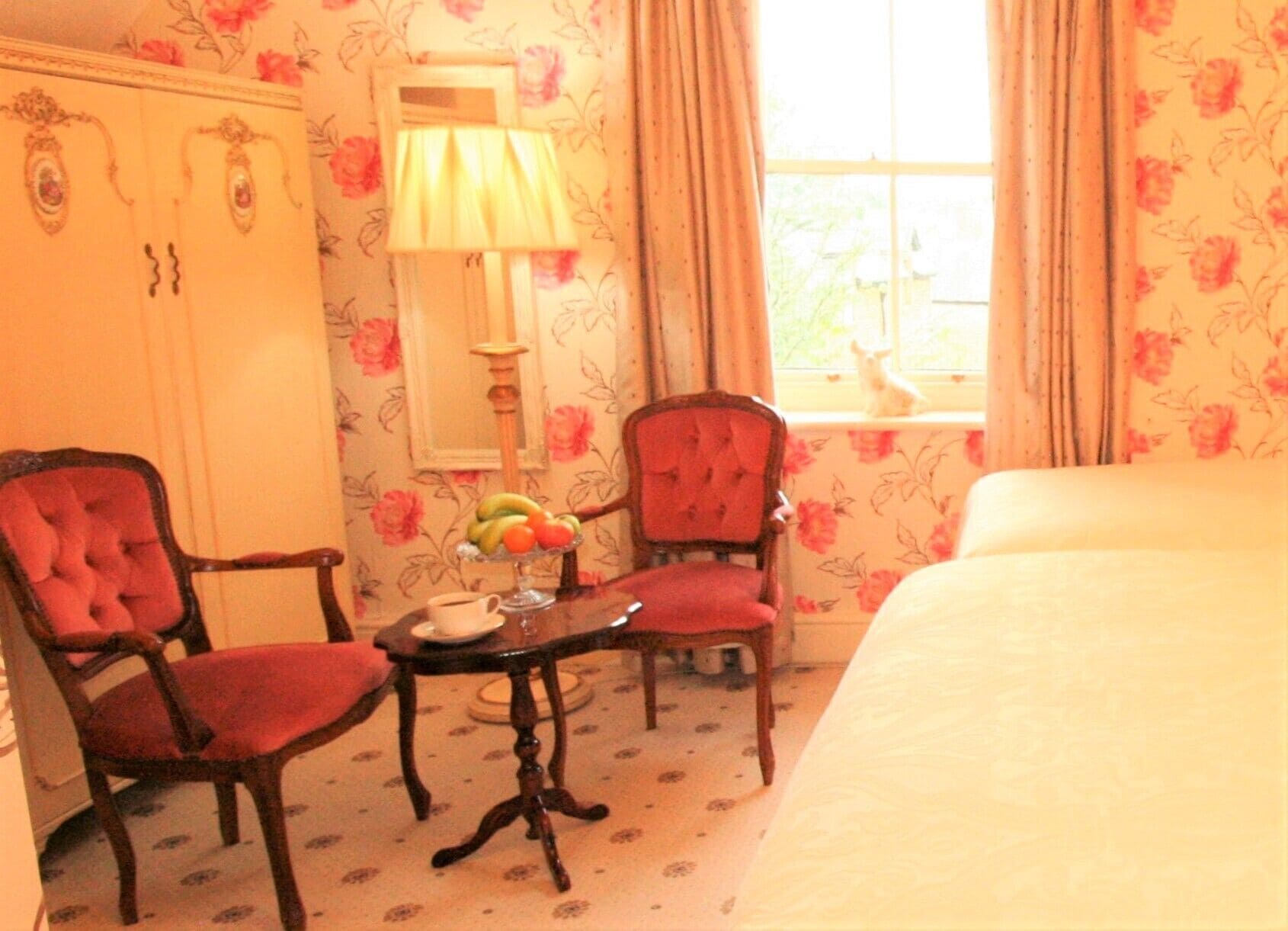 Habitación de hotel con cama, dos sillones tapizados en rojo, una mesita con frutero, una lámpara y una ventana con cortinas.