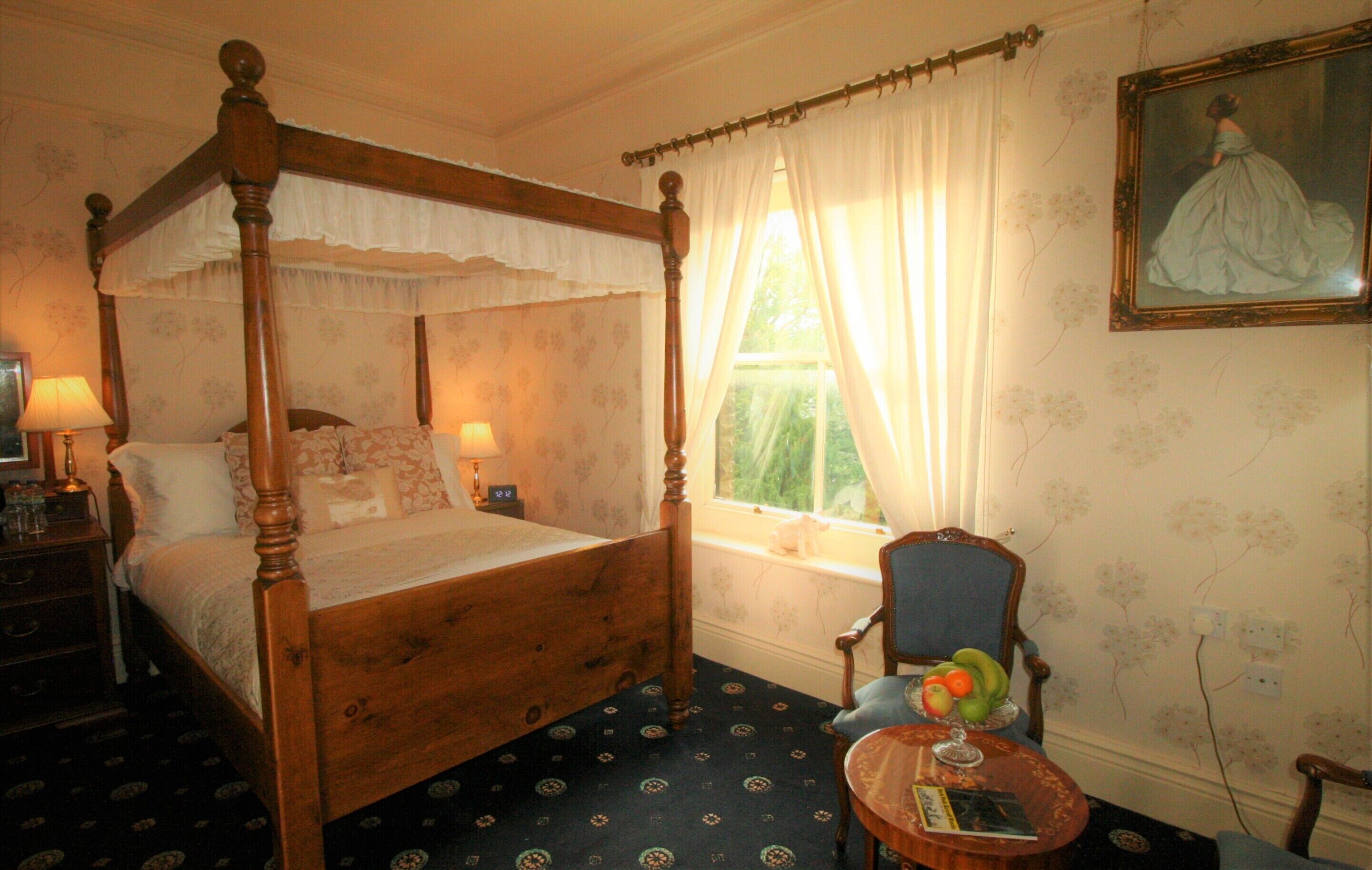 A four-poster bed with a canopy, a bedside table with a lamp, a chair, a small round table with a book and fruit, and a painting on the wall.