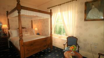 A four-poster bed with a canopy, a bedside table with a lamp, a chair, a small round table with a book and fruit, and a painting on the wall.