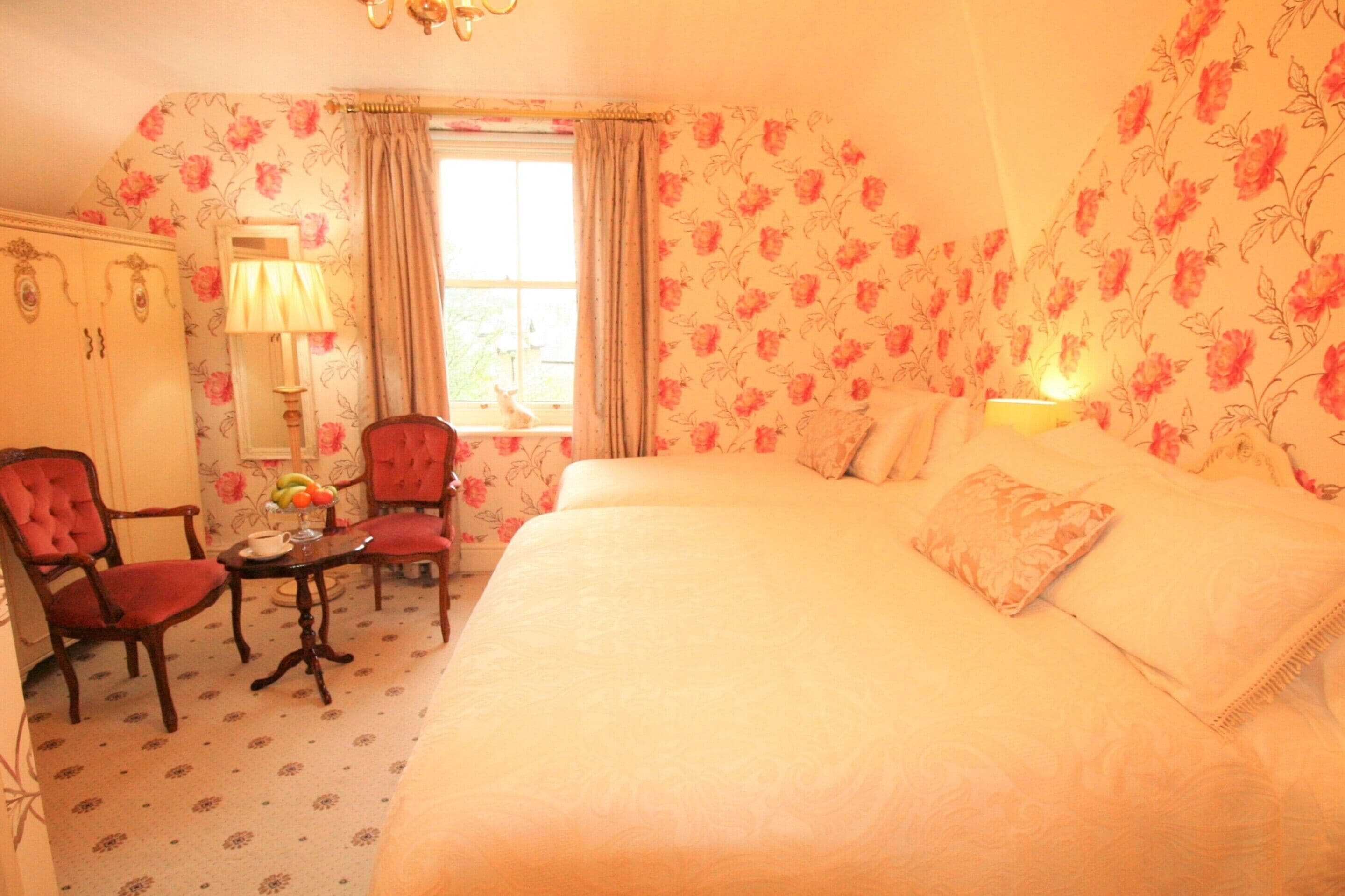 Un dormitorio con papel tapiz floral, una cama con almohadas, una mesita de noche con lámpara y dos sillas.