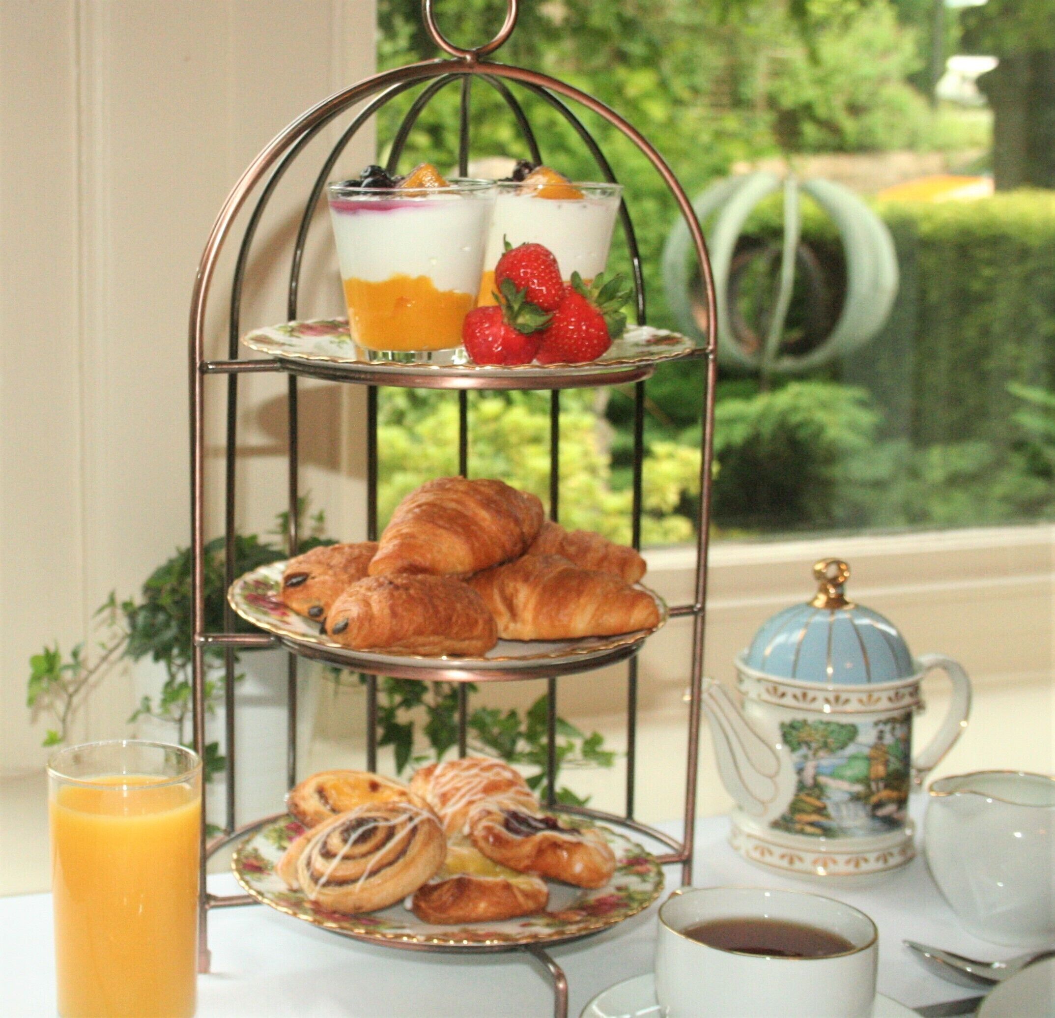 A three-tiered stand with pastries, a glass of orange juice, and a cup of coffee on a table by a window with a view of greenery.