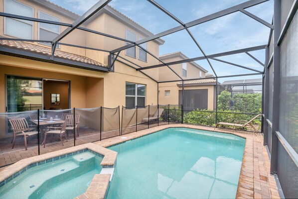 A swimming pool with a glass enclosure, a patio with chairs, and a view of neighboring buildings.