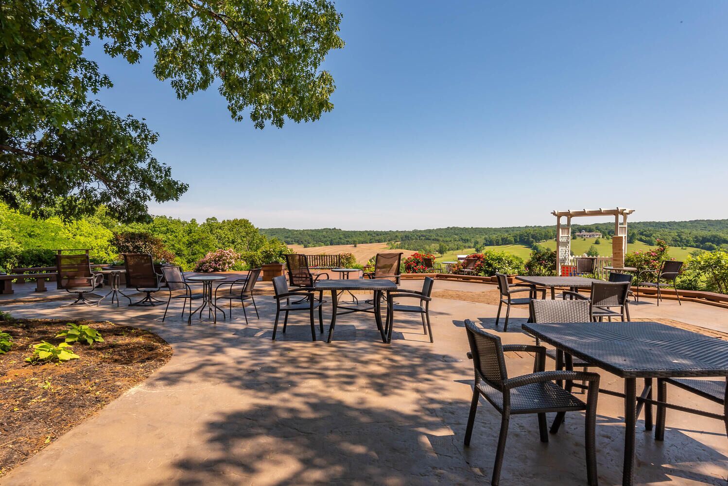 Un patio al aire libre con mesas y sillas, rodeado de vegetación y vistas a colinas ondulantes.